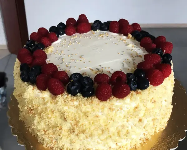 Pyszny tort śmietankowo-malinowy z mascarpone, ozdobiony świeżymi malinami i borówkami, posypany złotymi i srebrnymi kuleczkami.
