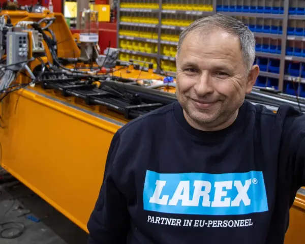 Uśmiechnięty pracownik w bluzie z logo Larex Deventer stoi obok pomarańczowej maszyny w warsztacie.