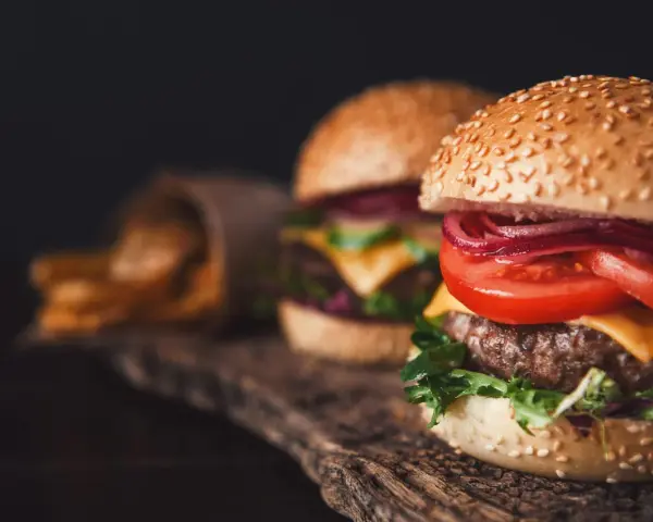 Dwa soczyste burgery z serem, pomidorem i czerwoną cebulą na drewnianej desce, obok frytki.