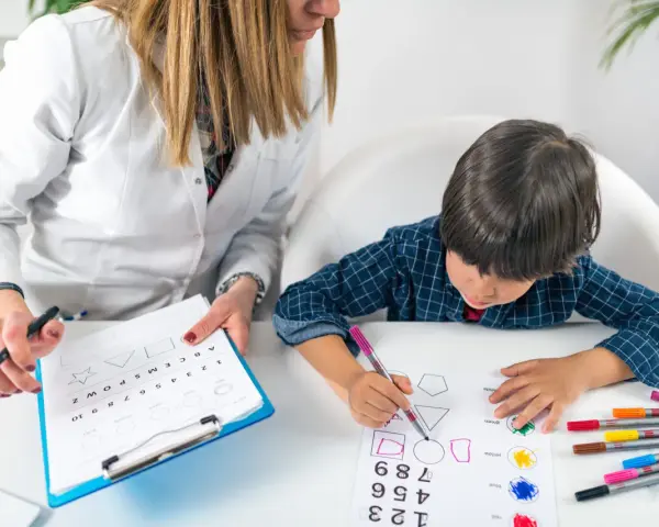 Bambino impegnato in un test per l'autismo, colora forme geometriche guidato da un'insegnante.