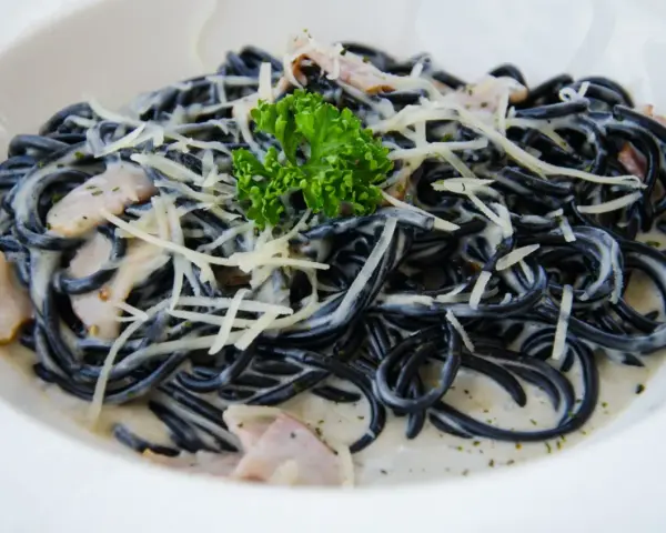Czarny makaron spaghetti w kremowym sosie, posypany startym parmezanem i udekorowany gałązką natki pietruszki.