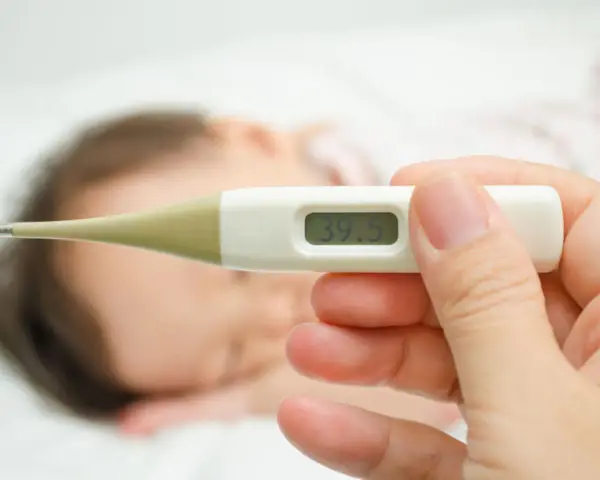 Cyfrowy termometr pokazuje 39.5°C. W tle widać śpiące dziecko. Zastanawiasz się, ile czasu mierzyć temperaturę pod pachą?
