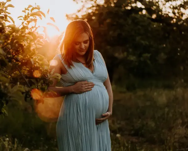 Piękne zdjęcia w ciąży. Kobieta w błękitnej sukience trzyma dłonie na brzuchu, otoczona ciepłym światłem zachodzącego słońca.
