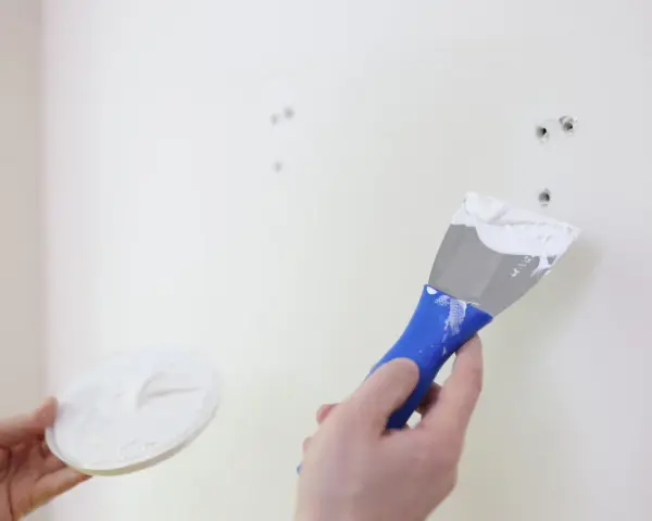Persona aplica masilla en una pared blanca con una espátula azul para reparar agujeros.