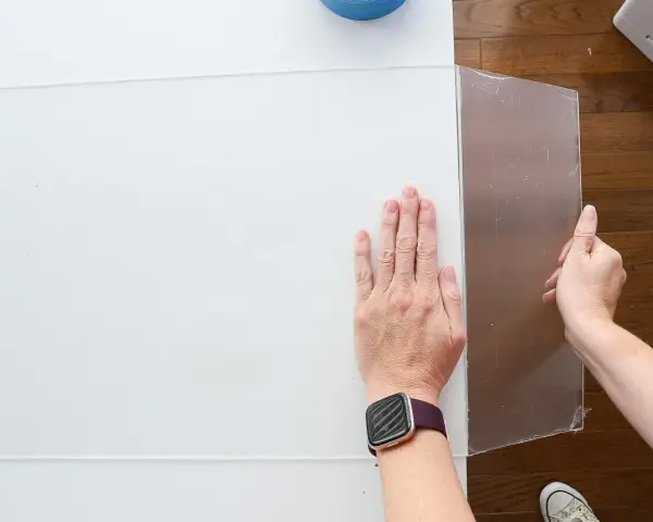 Hands guide a scoring tool along a white acrylic sheet, demonstrating how to cut acrylic sheet for a project.