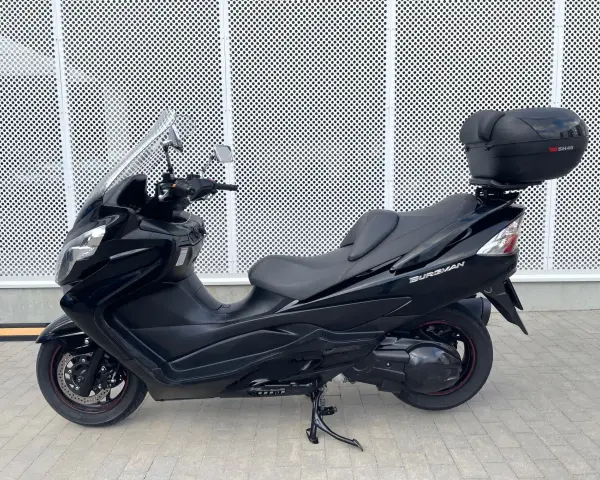 Czarny skuter Suzuki Burgman 400 z kufrem SH46 na tle perforowanej białej ściany.