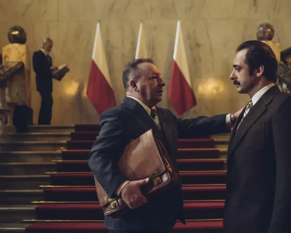 Hubert Niedziela, w garniturze, trzyma teczkę, rozmawiając z mężczyzną na schodach przed polskimi flagami.