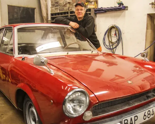 Man lutar sig mot en röd klassisk bil i garage. Lite rost syns på bilens skärmkanter.