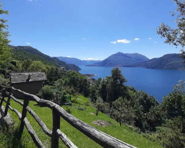 Sentiero del Viandante tappe: un casolare in pietra con tetto in ardesia e un recinto in legno rustico si affacciano su un lago blu intenso circondato da montagne verdi.