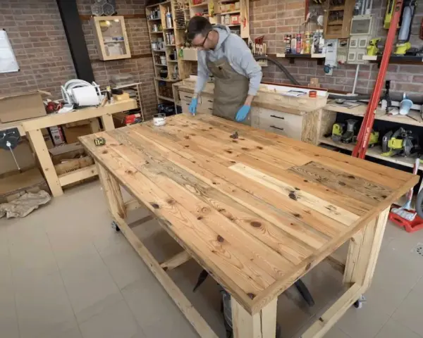 Uomo con grembiule lavora su un banco da lavoro con pallet, applicando finitura al legno. L'officina è piena di attrezzi e materiali.