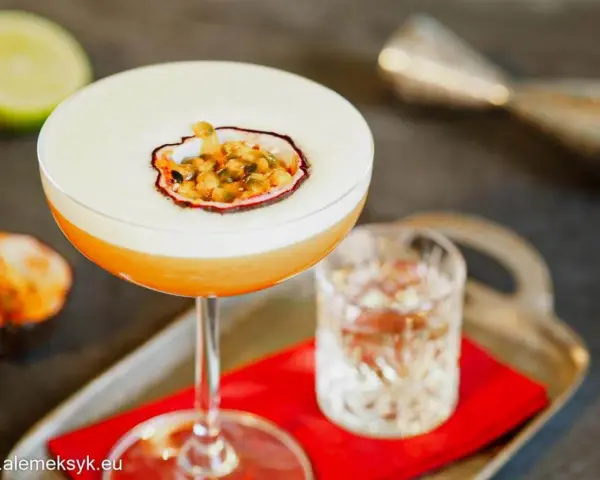Orzeźwiający drink z martini, ozdobiony połówką marakui, obok kieliszka z wodą i połówki limonki.