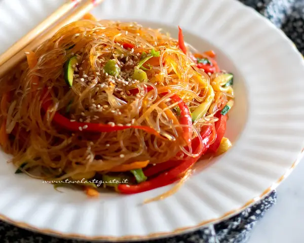Un piatto di spaghetti di soia con verdure colorate, peperoni rossi, zucchine e carote, guarnito con semi di sesamo.