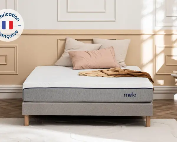 Matelas gris et blanc avec logo "mello", sur un sommier, avec des coussins et un plaid.