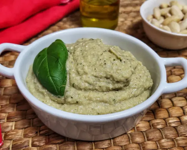 Pesto di melanzane cremoso in una ciotola bianca, guarnito con una foglia di basilico. Sullo sfondo, mandorle e olio d'oliva.