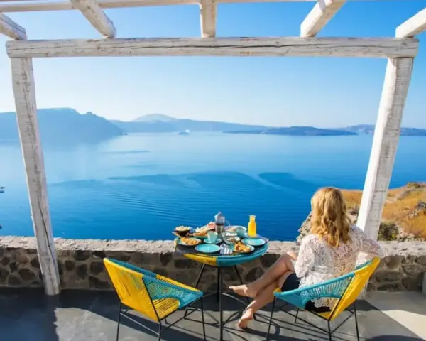 Mujer desayunando en balcón con vista al mar azul y montañas.