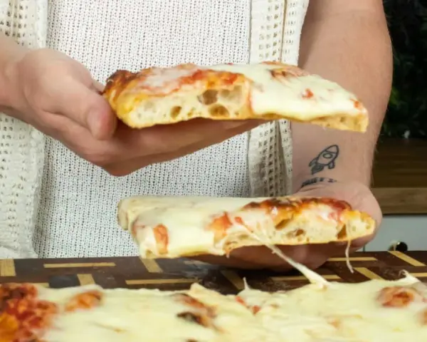 Due fette di pizza in teglia, con mozzarella filante e crosta dorata, vengono sollevate da mani.