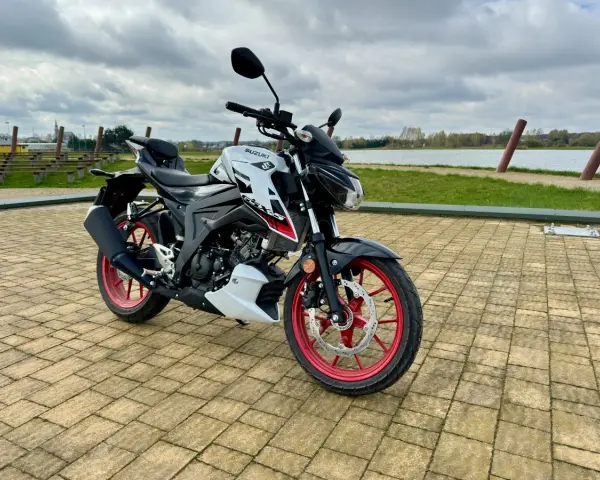 Biało-czarny Suzuki GSX S125 z czerwonymi felgami stoi na wybrukowanym placu nad jeziorem.