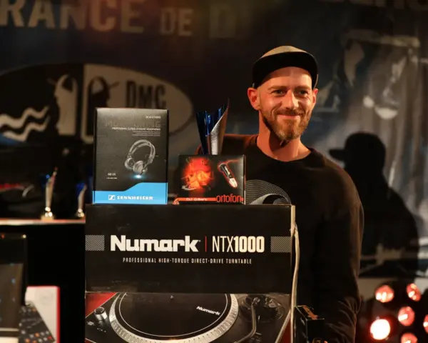 Un DJ souriant pose avec des prix et du matériel, dont des casques Sennheiser et un tourne-disque Numark. Une liste DJ internationale est visible.