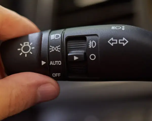 Dłoń reguluje przełącznik świateł auto. Widoczne symbole oznaczają światła drogowe, mijania, postojowe i automatyczne.