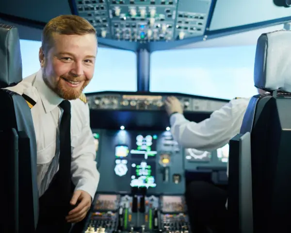 Uśmiechnięty pilot w kabinie samolotu, obok drugiego pilota przy panelu sterowania.