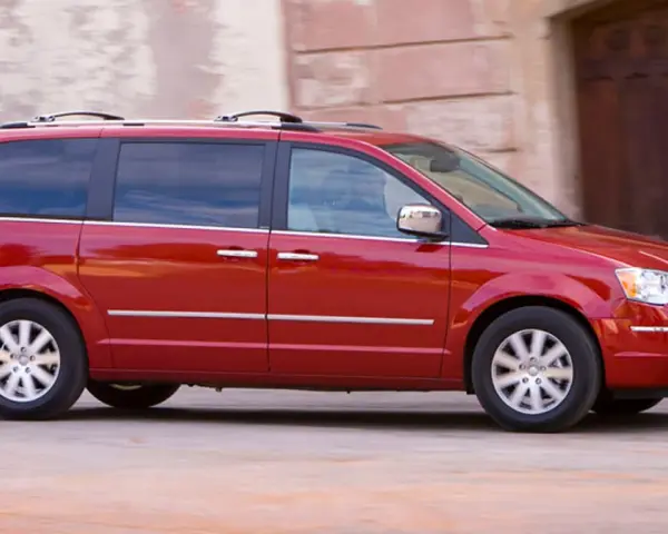 Czerwony Chrysler Grand Voyager w ruchu, z przyciemnianymi szybami i relingami dachowymi, przemierza ulicę obok kamiennej fasady.