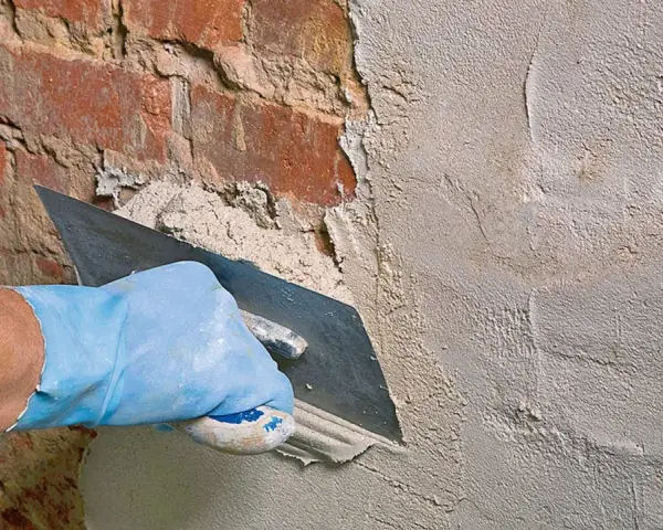 Hand mit blauer Handschuh glättet mit Kelle den **Sanierputz Keller** auf rotem Ziegelmauerwerk.