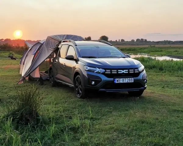 Dacia Jogger Camper rozłożony na biwaku o zachodzie słońca. Namiot połączony z autem, otoczony łąką i wodą.