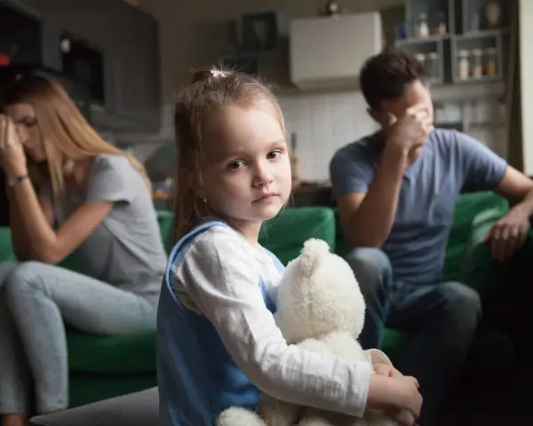 Smutna dziewczynka z misiem w objęciach, podczas gdy rodzice w tle przeżywają trudne chwile. Temat: rozwód a dzieci.