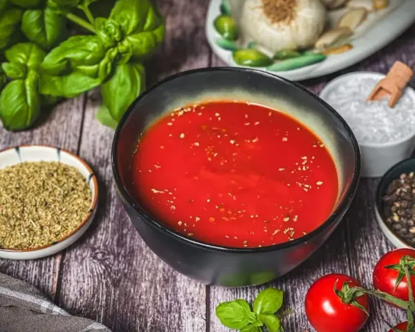 Schüssel mit leuchtend roter Pizza Sauce, umgeben von frischem Basilikum, Tomaten, Oregano, Salz und Pfeffer – wie in der Pizzeria.