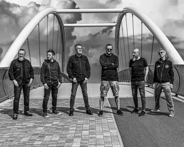 Sześciu mężczyzn, zespół Strachy na Lachy, pozuje na moście. Gotowi na koncerty, w rockowym stylu, pod burzliwym niebem.