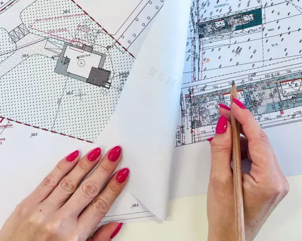 Dłoń z różowym lakierem na paznokciach trzymająca ołówek nad mapą projektową.