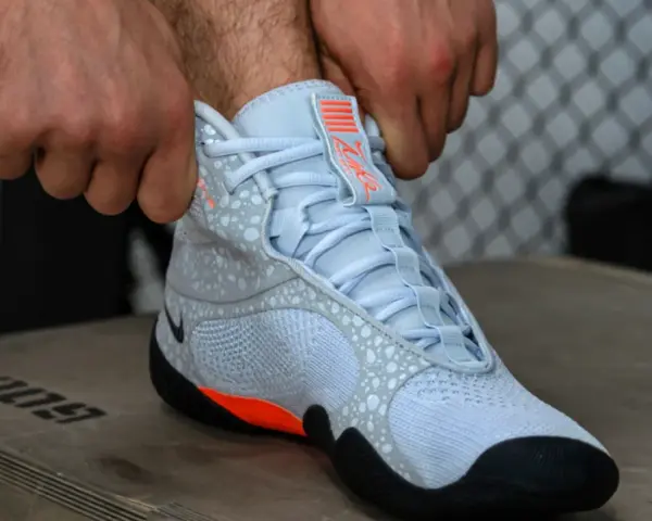Ręce zapinają jasnoniebieskie buty sportowe z pomarańczowymi akcentami, przypominające styl Batistini.