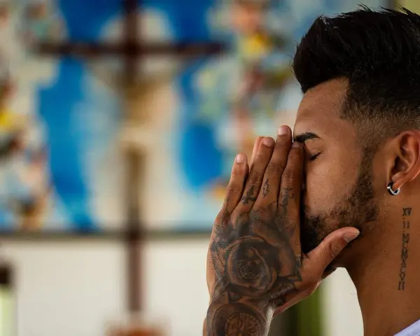 A man with tattoos prays before a crucifix, contemplating if is vaping a sin.