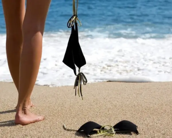 Nogi na piaszczystej plaży, obok czarny kostium kąpielowy. Fale obmywają brzeg. Idealne miejsce na naturystyczną plażę.