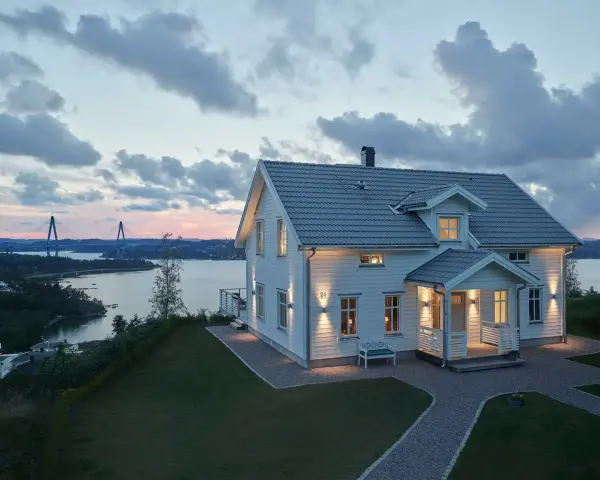 Vackert vitt hus med upplysta fönster och entré, perfekt hus entre inspiration. Vacker vy över vatten och bro i skymningen.
