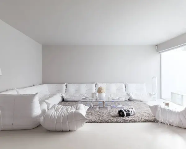 Soggiorno minimalista con divano componibile bianco, tappeto grigio e tavolino trasparente. Le tonalità di bianco per pareti creano un'atmosfera eterea.