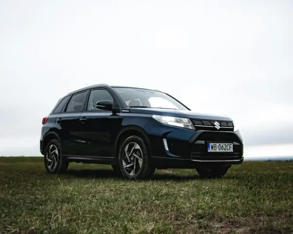 Ciemnoniebieski Suzuki Vitara Hybrid na tle zachmurzonego nieba, zaparkowany na trawiastym wzgórzu.