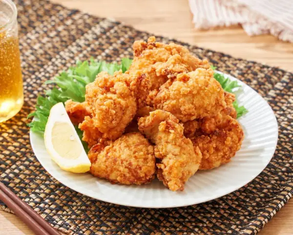 Chrupiące karaage chicken na talerzu, ozdobione plasterkiem cytryny i liśćmi sałaty. Obok szklanka złocistego napoju i pałeczki.