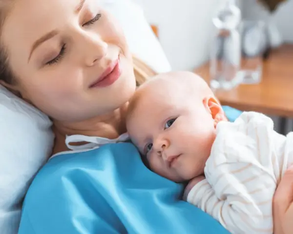 Uśmiechnięta mama tuli noworodka. To chwila spokoju po szczepieniach noworodka, budująca więź i zapewniająca zdrowie maluszka.