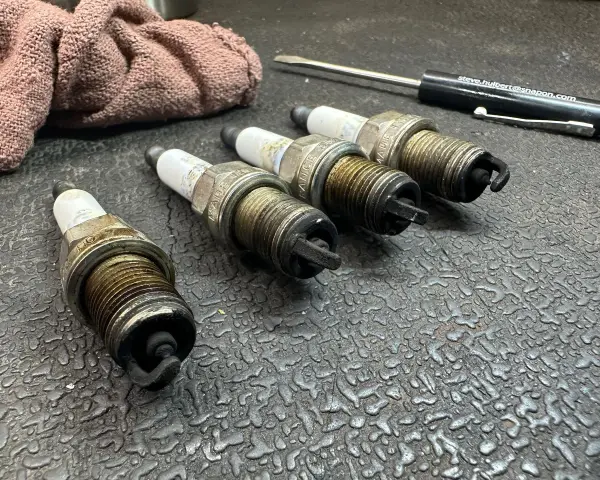 Four used spark plugs lie on a textured surface next to a rag and a screwdriver. This image might make you wonder how many spark plugs a car has.