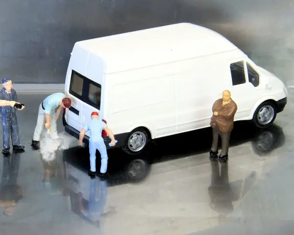 Miniaturowi pracownicy zajmują się białym busem budowlanym. Jeden z nich czyści tył pojazdu, inny trzyma narzędzie, a trzeci obserwuje.