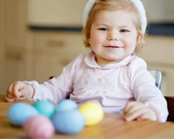 Kleines Mädchen mit Hasenohren und bunten Ostereiern. Sie sitzt am Tisch und schaut neugierig auf das **Baby Ei**.