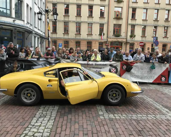 Żółte Ferrari Dino z otwartymi drzwiami, kierowca w środku, tłum widzów za barierką.