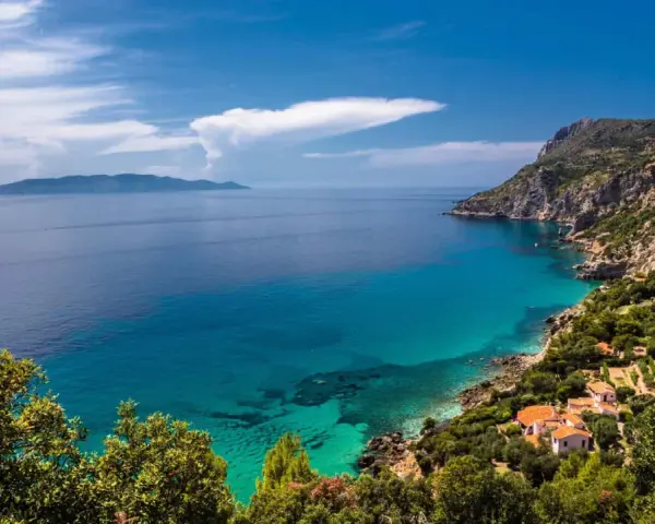 Turquoise water laps a rocky Tuscan coast with villas nestled among trees, under a blue sky with clouds.
