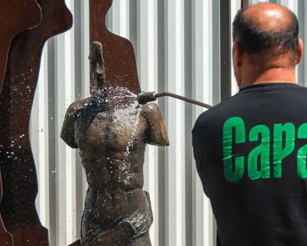 Hombre con camiseta negra "Capa" limpia una escultura de bronce con agua a presión.