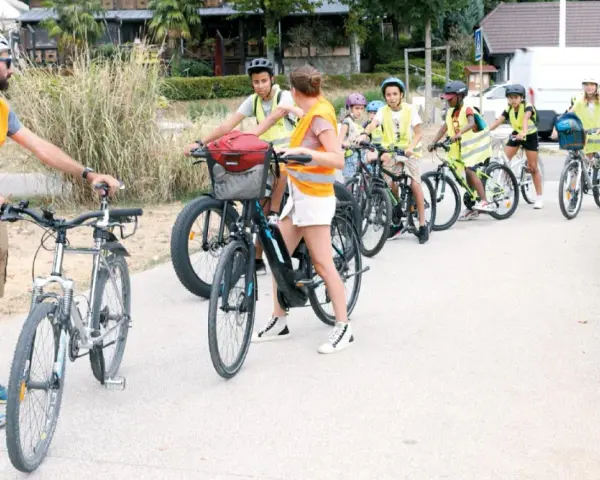 Un groupe d'enfants à vélo, encadrés par des adultes en gilets de sécurité, suit un parcours. La scène illustre la réglementation du déplacement à pied et à vélo à l'ACM.
