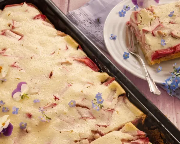 Saftiger Rhabarberkuchen mit Quark, dekoriert mit Veilchen und Vergissmeinnicht. Ein Stück ist auf einem Teller angerichtet.