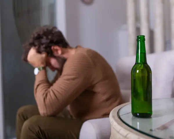 Mężczyzna siedzi z głową w dłoniach, obok stoi zielona butelka. Czuje duszności po alkoholu.