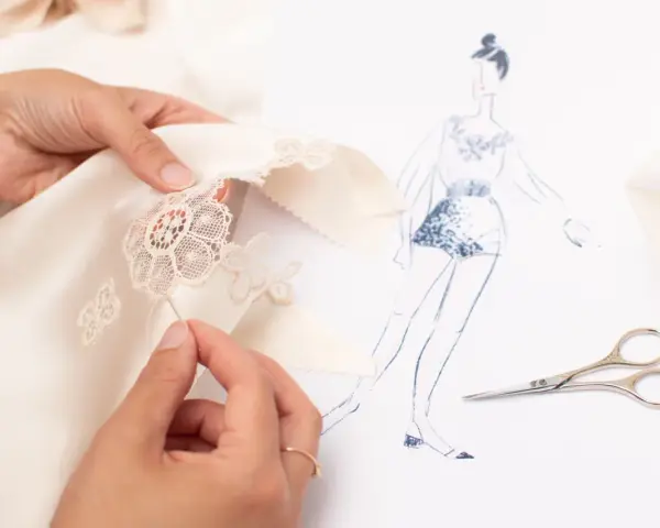 Main d'une couturière réalisant un assemblage couture délicat sur un tissu de soie, avec un croquis de mode et des ciseaux à proximité.