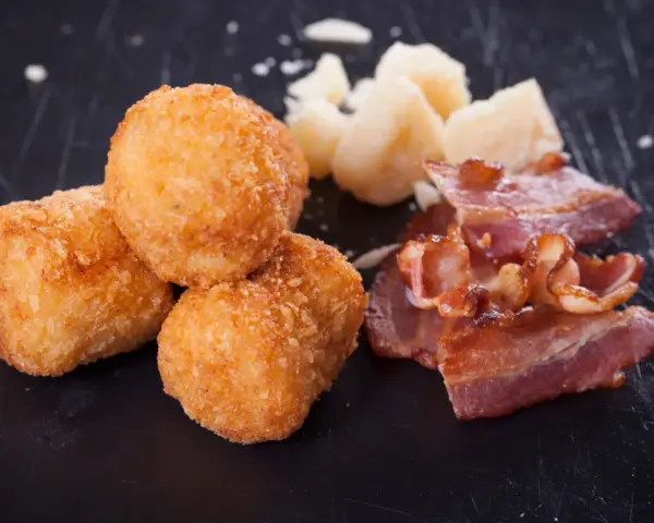 Croquetas doradas y crujientes con trozos de bacon y queso sobre fondo oscuro.